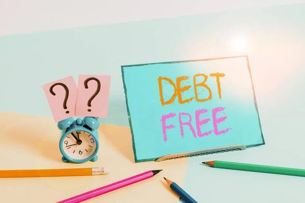 Writing note showing Debt Free. Business photo showcasing free from owning any money to any individual or a company Mini size alarm clock beside stationary on pastel backdrop.