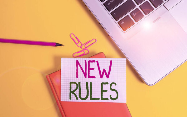 Writing note showing New Rules. Business photo showcasing recently one of a set of explicit or understood regulations Laptop clips pencil paper sheet hard cover notebook colored background.