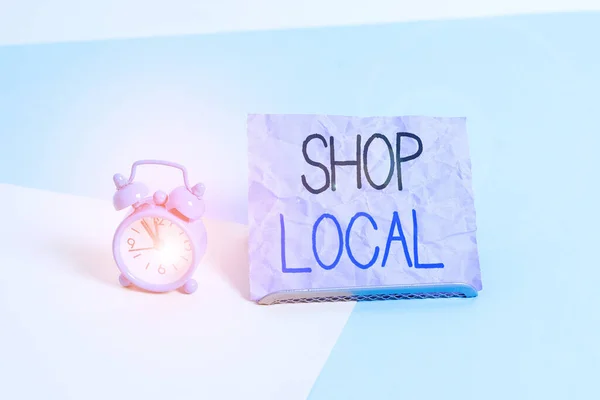 Handwriting text writing Shop Local. Concept meaning a preference to buy locally produced goods and services Mini size alarm clock beside a Paper sheet placed tilted on pastel backdrop.