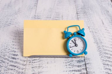 Mini mavi alarm saati sarı bir not kağıdının önüne yerleştirilmiş zamanı gösterir. Simge kablosunun üzerinde duran klasik küçük saat gösterim kağıdının önünde