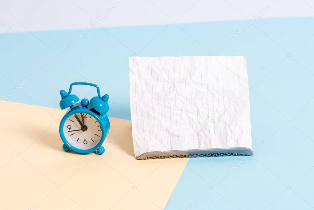 Mini small size alarm clock beside a Paper sheet placed tilted. Notepaper on the edge of empty plain multi colours table backdrop. Artistic way of arranging flat lays photography