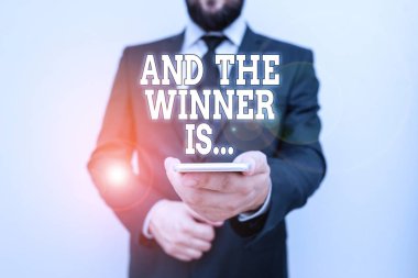 Text sign showing And The Winner Is. Conceptual photo Announcing who the Victor is Exemplar Achiever Male human wear formal work suit hold smart hi tech smartphone use one hand. clipart