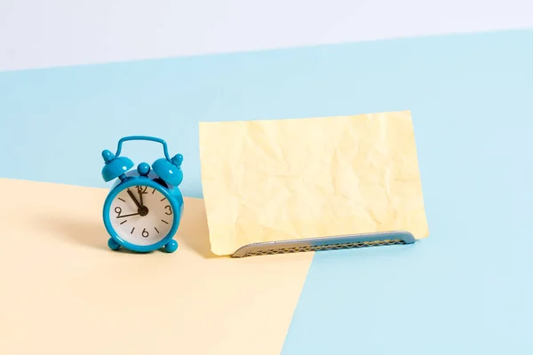 Mini klein formaat wekker naast een papieren blad geplaatst gekanteld. Notepaper op de rand van lege effen multi kleuren tafel achtergrond. Artistieke manier om plat te leggen fotografie — Stockfoto