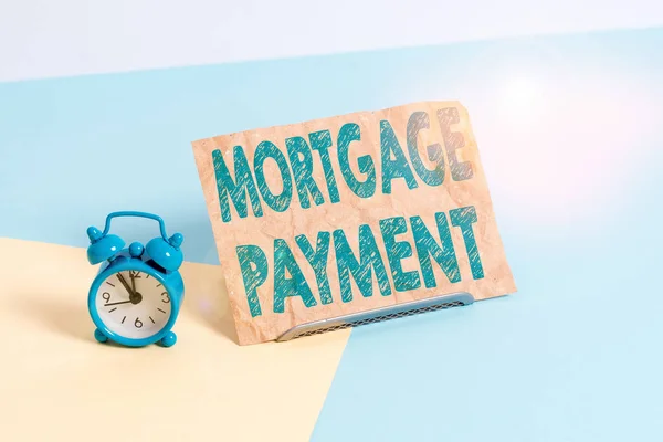 Text sign showing Mortgage Payment. Conceptual photo periodic amount paid to a holder for repayment of a loan Mini size alarm clock beside a Paper sheet placed tilted on pastel backdrop.