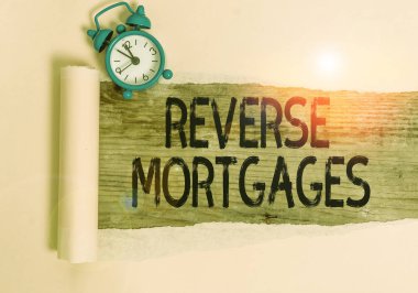 Text sign showing Reverse Mortgages. Conceptual photo borrower to access the unencumbered value of the property Alarm clock and torn cardboard placed above a wooden classic table backdrop. clipart