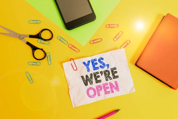 Text sign showing Yes We Re Open. Conceptual photo answering on client that shop is available at this time Paper sheets pencil clips smartphone scissors notebook colored background. — Stockfoto