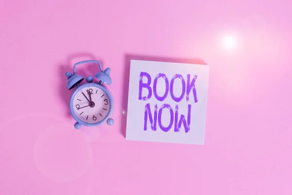 Writing note showing Book Now. Business photo showcasing Make a reservation in hotel accommodation or airplane ticket Vintage alarm clock wakeup blank notepad sticky note colored background.