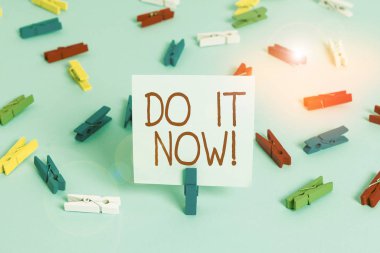 Do It Now 'ı gösteren bir not yazıyorum. İş fotoğrafı gösterimi tereddüt etmeden çalışmaya başlayın ya da hemen bir şeyler yapın. Renkli çamaşır iğnesi kağıtları boş mavi zemin ofisini anımsatıyor..