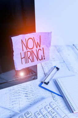 Writing note showing Now Hiring. Business photo showcasing finding evaluating working relationship with future employees Note paper taped to black computer screen near keyboard and stationary.