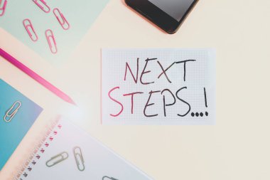 Sonraki Adımları gösteren bir not yazıyorum. İş fotoğrafında ilkini bitirdikten sonra yaptığın bir şeyi gösteriyorsun. Kağıt kağıtlar kare kalem tokaları, akıllı telefon renkli arka plan..