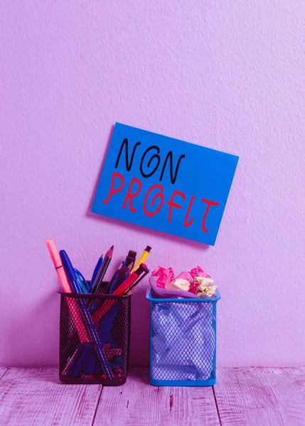 Text sign showing Non Profit. Business photo text type of organization that does not earn profits for its owners Blue Sticky Card on Wall Two Pencil Pots Pens Pencils Markers Waste Paper