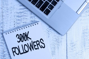 300.000 takipçiyi gösteren bir not yazıyorum. İş fotoğraflarında Instagram 'da birini takip eden kişi sayısı gösteriliyor. Üst düzey metalik dizüstü bilgisayar boş spiral not defteri ahşap masada yatıyor..