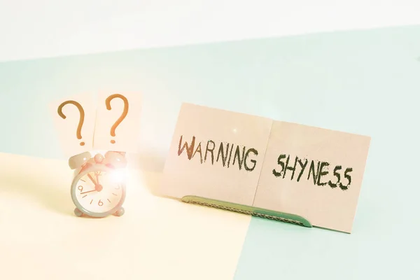 Word writing text Warning Shyness. Business concept for Lack of confidence Not able to speak out or express oneself Mini size alarm clock beside a Paper sheet placed tilted on pastel backdrop.