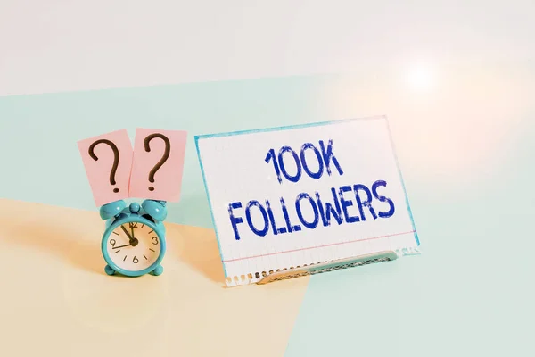 Segno di testo che mostra 100K seguaci. Concettuale numero di foto di persone che seguono qualcuno in Instagram Mini dimensione sveglia accanto a un foglio di carta posto inclinato su sfondo pastello . — Foto Stock