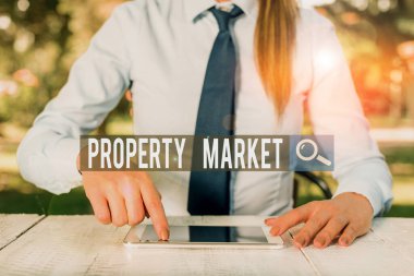 Writing note showing Property Market. Business photo showcasing the buying and selling of land and buildings Estate market Female business person sitting by table and holding mobile phone.