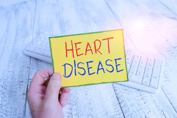 Handwriting text Heart Disease. Concept meaning class of diseases that involve the heart or blood vessels man holding colorful reminder square shaped paper white keyboard wood floor. — Zdjęcie stockowe