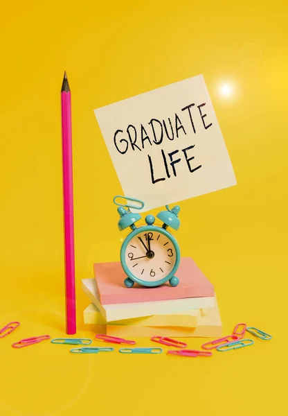 Writing note showing Graduate Life. Business photo showcasing condition or a status a demonstrating after finishing academic degree Alarm clock sticky note stacked notepads pencil colored background. — Stockfoto