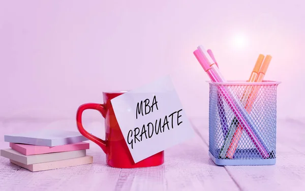 Handwriting text writing Mba Graduate. Concept meaning masters is degree with specialization in business administration Coffee cup blank sticky note pens metal holder stacked pads wooden table.