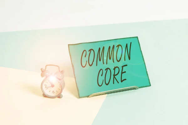 Conceptual hand writing showing Common Core. Business photo showcasing set of academic standards in mathematics and English language Alarm clock beside a Paper sheet placed on pastel backdrop.