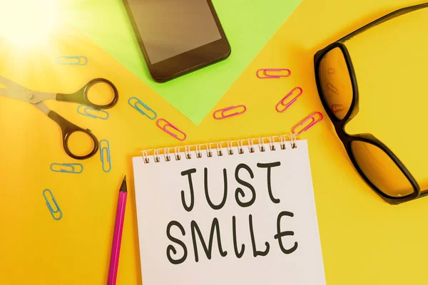 Scrivere una nota che mostra Just Smile. Business photo showcase assumere un'espressione facciale che indica piacere o divertimento Foglio matita smartphone forbici occhiali blocco note colore sfondo . — Foto Stock