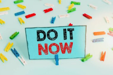 Do It Now 'ı gösteren bir not yazıyorum. İş fotoğrafı gösterimi tereddüt etmeden çalışmaya başlayın ya da hemen bir şeyler yapın. Renkli çamaşır iğnesi kağıtları boş mavi zemin ofisini anımsatıyor..