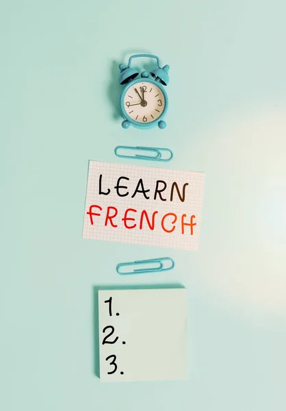 Text sign showing Learn French. Conceptual photo get knowledge or skill in speaking and writing French language Alarm clock squared blank paper sheet notepad clips colored background.