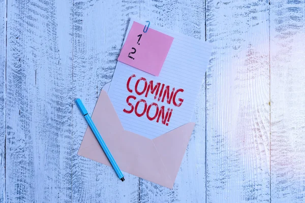 Text sign showing Coming Soon. Conceptual photo event or action that will happen after really short time Envelope blank sheet letter paper sticky note ballpoint wooden background.