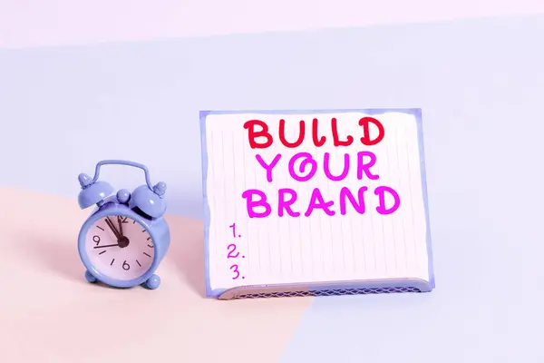 Writing note showing Build Your Brand. Business photo showcasing enhancing brand equity using advertising campaigns Alarm clock beside a Paper sheet placed on pastel backdrop. — Stock Photo, Image