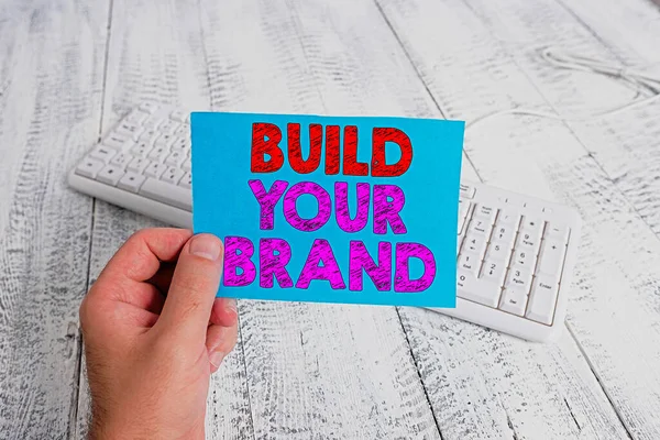 Handwriting text writing Build Your Brand. Concept meaning enhancing brand equity using advertising campaigns man holding colorful reminder square shaped paper white keyboard wood floor. — Stock Photo, Image