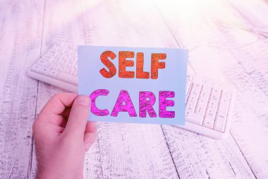 Kişisel Bakım 'ın el yazısı. Kavram, birini geliştirmek için harekete geçme uygulaması elinde renkli kare şeklinde kağıt beyaz klavye ahşap zemin tutan kendi sağlıkçısıdır..