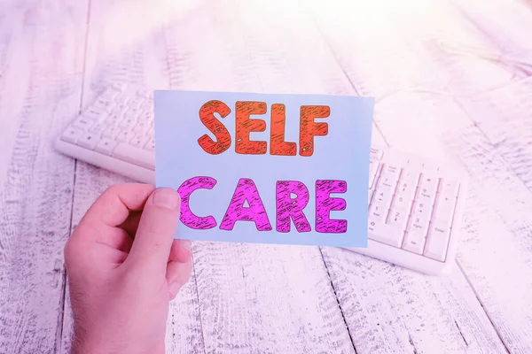 Escrita à mão escrita de texto Self Care. Conceito que significa a prática de tomar medidas para melhorar um é o próprio homem de saúde segurando lembrete colorido quadrado em forma de papel branco teclado piso de madeira . — Fotografia de Stock