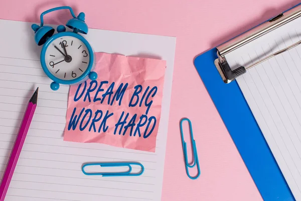 Text sign showing Dream Big Work Hard. Conceptual photo Believe in yourself and follow the dreams and goals Paper sheet note clipboard pencil clips alarm clock colored background.