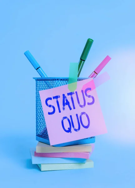 Text sign showing Status Quo. Conceptual photo existing state of affairs regarding social or political issues Sticky note arrow banners stacked pads metal pens holder pastel background.