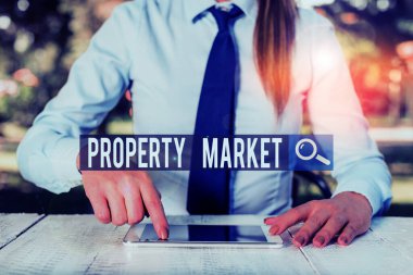 Writing note showing Property Market. Business photo showcasing the buying and selling of land and buildings Estate market Female business person sitting by table and holding mobile phone.