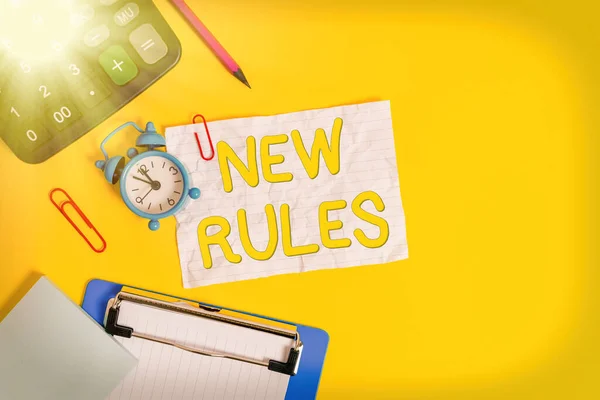 Writing note showing New Rules. Business photo showcasing recently one of a set of explicit or understood regulations Alarm clock crushed note calculator pencil clipboard color background.