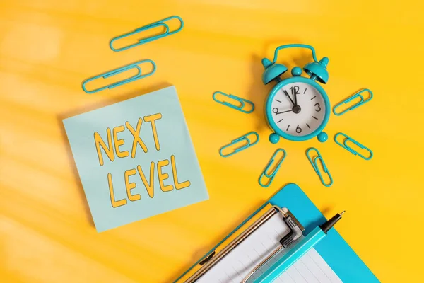 Handschrift tekst schrijven Next Level. Concept betekent dat het hebben van meer dan een verdieping en verwijzend naar na de huidige metalen wekker klembord clips balpen notitieblok gekleurde achtergrond. — Stockfoto