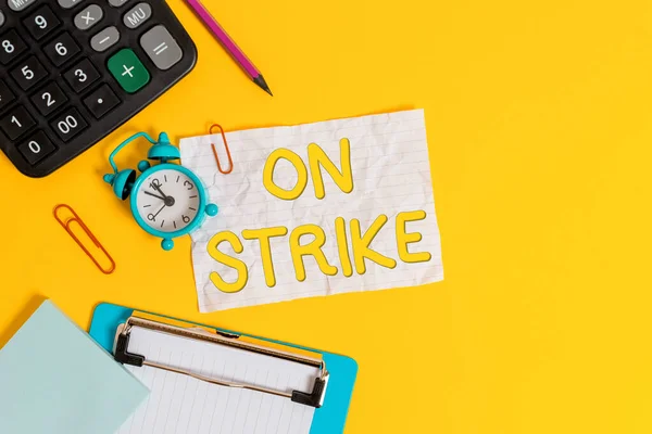 Writing note showing On Strike. Business photo showcasing refuse to continue working because of an argument with an employer Alarm clock crushed note calculator pencil clipboard color background.