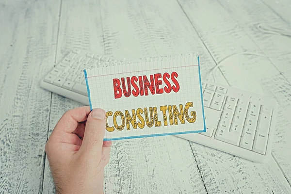 Text sign showing Business Consulting. Conceptual photo Blends Practice of Academic Theoretical Expertise man holding colorful reminder square shaped paper white keyboard wood floor.