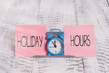 Holiday Hours 'u gösteren bir not yazıyorum. İş fotoğrafı gösterimi Programı 24 ya da 7 Yarım Gün Son Dakika Geç Kapanış Mini mavi alarm saati iki kağıt teli arasında tampon telin üzerinde duruyor.