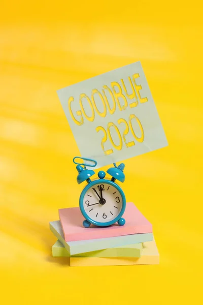 Handwriting text Good Bye 2020. Concept meaning express good wishes when parting or at the end of last year Metal alarm clock blank sticky note stacked notepads colored background.