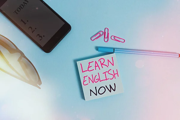 Word writing text Learn English Now. Business concept for gain or acquire knowledge and skill of english language Eyeglasses colored sticky note smartphone cell clips pen pastel background.