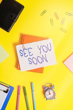See You Soon 'u gösteren bir not yazıyorum. Biriyle vedalaşmak ve tekrar buluşmaya gitmek için kullanılan iş fotoğrafı sergisi. Kalem ve ataçlarla dolu boş bir kağıdın üzerinde yatıyordu..