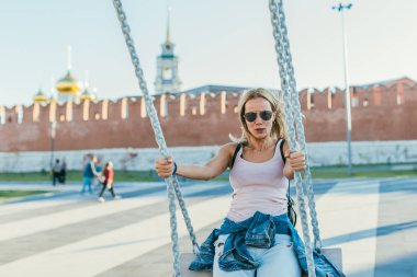 Uzun saçlı, pembe tişörtlü, güneş gözlüklü, açık mavi kot pantolonlu, salıncakta sallanan ve gülümseyen güzel bir sarışın. Sığ Dof ile seçici odak makro görüntüsü