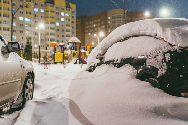 cars in the yard are completely covered with snow. In the background is Playground and residential buildings with glowing Windows. Quiet winter evening