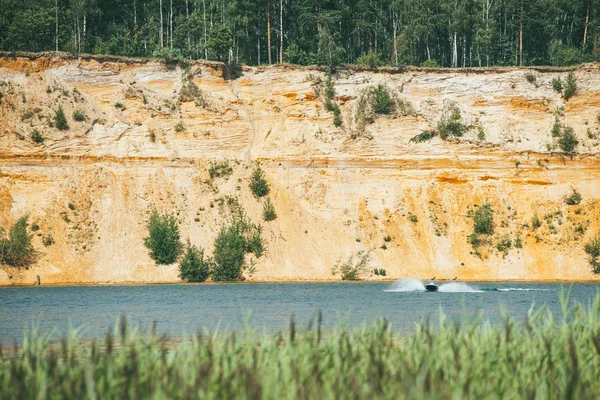Alta orilla empinada arenosa rocosa cubierta de pinos sobre un lago claro y azul. lancha rápida corta a través de la superficie del agua — Foto de Stock
