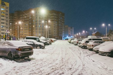 Bahçedeki arabalar tamamen karla kaplı. Arka planda oyun alanı ve parlayan pencereli konutlar var. Sessiz bir kış akşamı