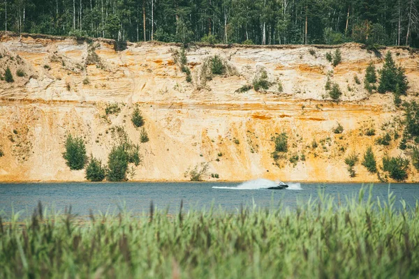Alta orilla empinada arenosa rocosa cubierta de pinos sobre un lago claro y azul. lancha rápida corta a través de la superficie del agua — Foto de Stock