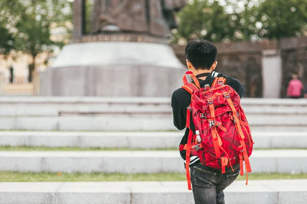 Um jovem fotógrafo asiático com uma mochila brilhante nos ombros fotografa o monumento. Vista traseira — Fotografia de Stock