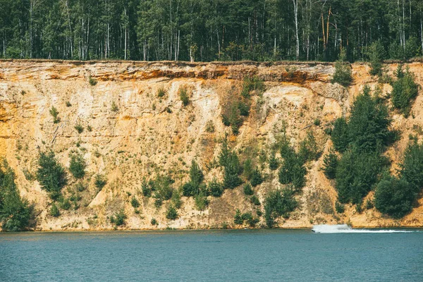 Alta orilla empinada arenosa rocosa cubierta de pinos sobre un lago claro y azul. lancha rápida corta a través de la superficie del agua — Foto de Stock