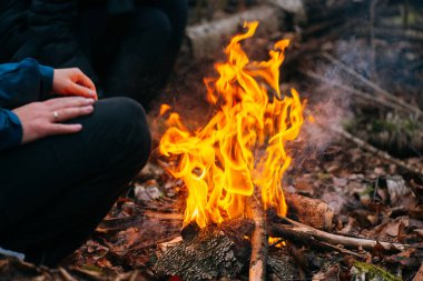 Adam ellerini ateşte ısıtıyor. Akşamları ormanda odun yakmak. Doğadaki turistik kampta kamp ateşi. Barbekü ve açık havada temiz hava. Ateş ve alev soyut karanlık arkaplanda kıvılcımlar saçar